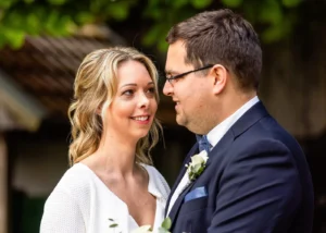 Brautpaar bei Hochzeitsfotografie – Mann in dunklem Anzug mit Brille und Fliege, Frau in weißem Kleid mit Blumenschmuck
