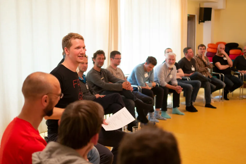 Gruppe von Männern in Seminarraum, ein Mann in schwarzem Shirt spricht zu der Gruppe