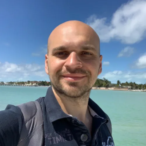 Konstantin, Mann mit Glatze am Strand vor türkisem Meer und blauem Himmel