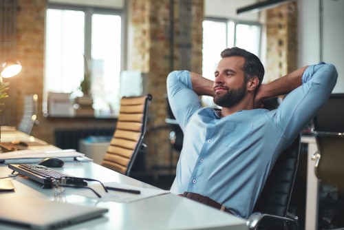 Mann in blauem Hemd sitzt am Schreibtisch, Hände hinter Kopf, relaxte Pose, modernes Büro
