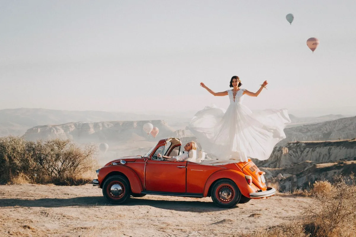 Braut in weißem Kleid mit ausgebreiteten Armen auf rotem Vintage-Cabrio in bergiger Landschaft mit Heißluftballons