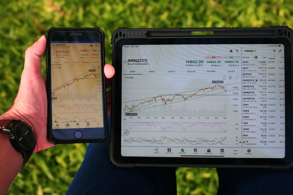 Smartphone und Tablet in Hand mit Handelsplattform und Aktiencharts, grüner Hintergrund mit Grasnarbe