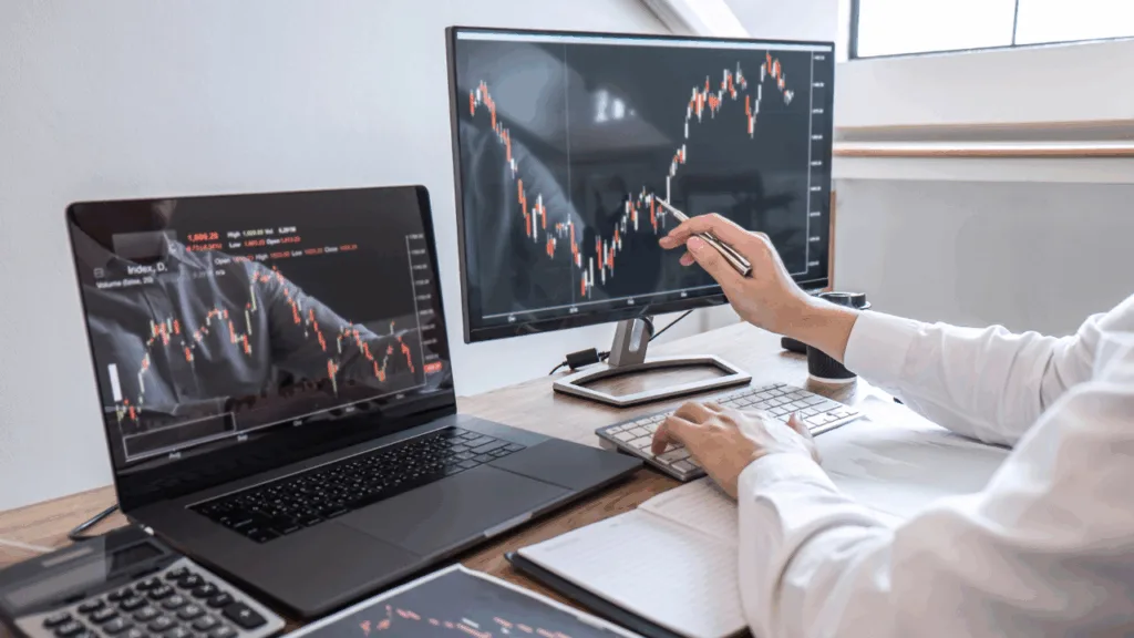 Person analysiert Börsendiagramme auf mehreren Bildschirmen, Laptop und Monitor mit Candlestick Charts