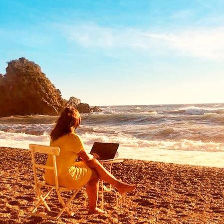 Rote Haare Frau sitzt am Strand vor Laptop, Meer und Felsen im Hintergrund