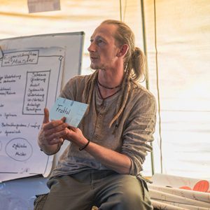 Mann mit blondem Haar vor beschriebenem Flipchart präsentiert Notizblock mit Permakultur-Inhalten