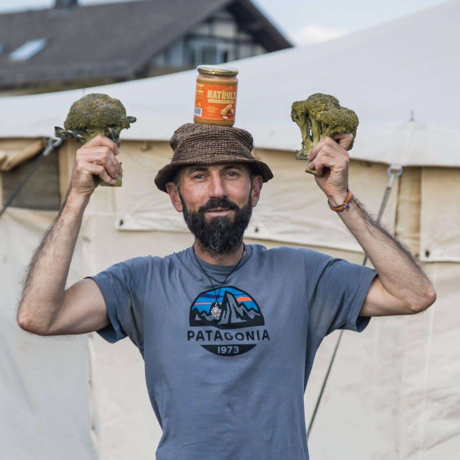 Mann mit Bart und Patagonia-Shirt hält zwei Brokkoli-Pflanzen und Naturreis-Glas humorvoll in die Höhe