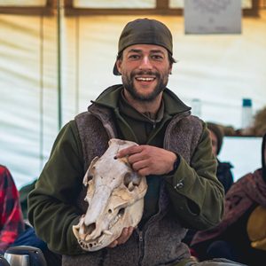 Mann mit dunklem Mütze präsentiert Tierschädel, lächelnd im Seminar-Setting