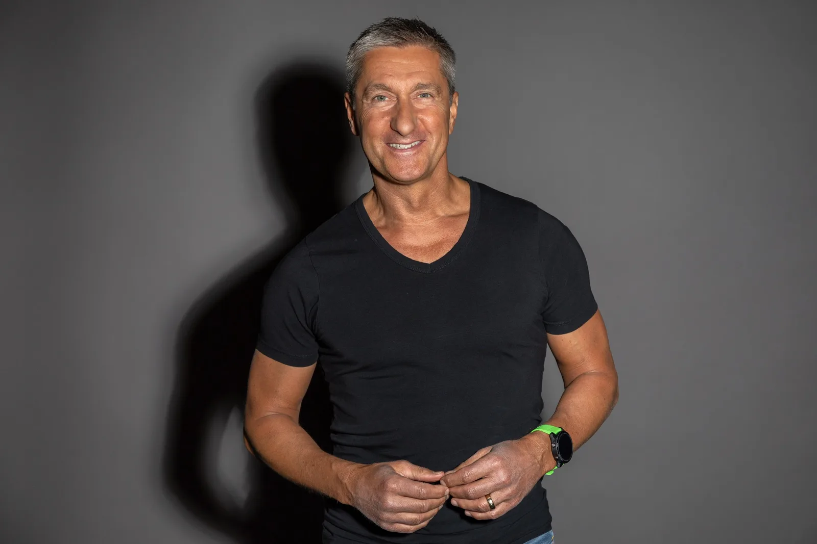 Alexander S. Kaufmann lächelt in die Kamera, trägt schwarzes T-Shirt und Smartwatch, porträtiert vor grauem Hintergrund