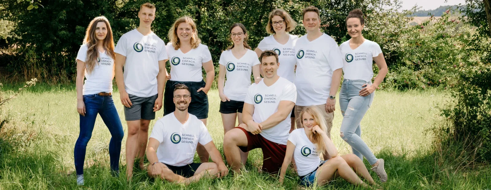 Team von neun Personen in weißen Shirts steht vor Hecke auf Wiese
