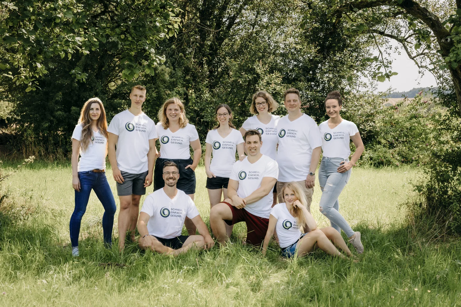 Team von neun Personen in weißen Shirts steht vor Baumhecke auf Wiese