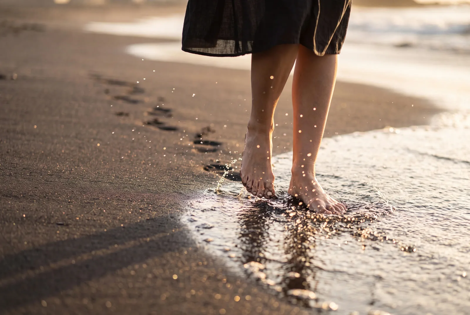 Barfüße waten durch glitzerndes Wasser am schwarzen Sandstrand im Abendlicht