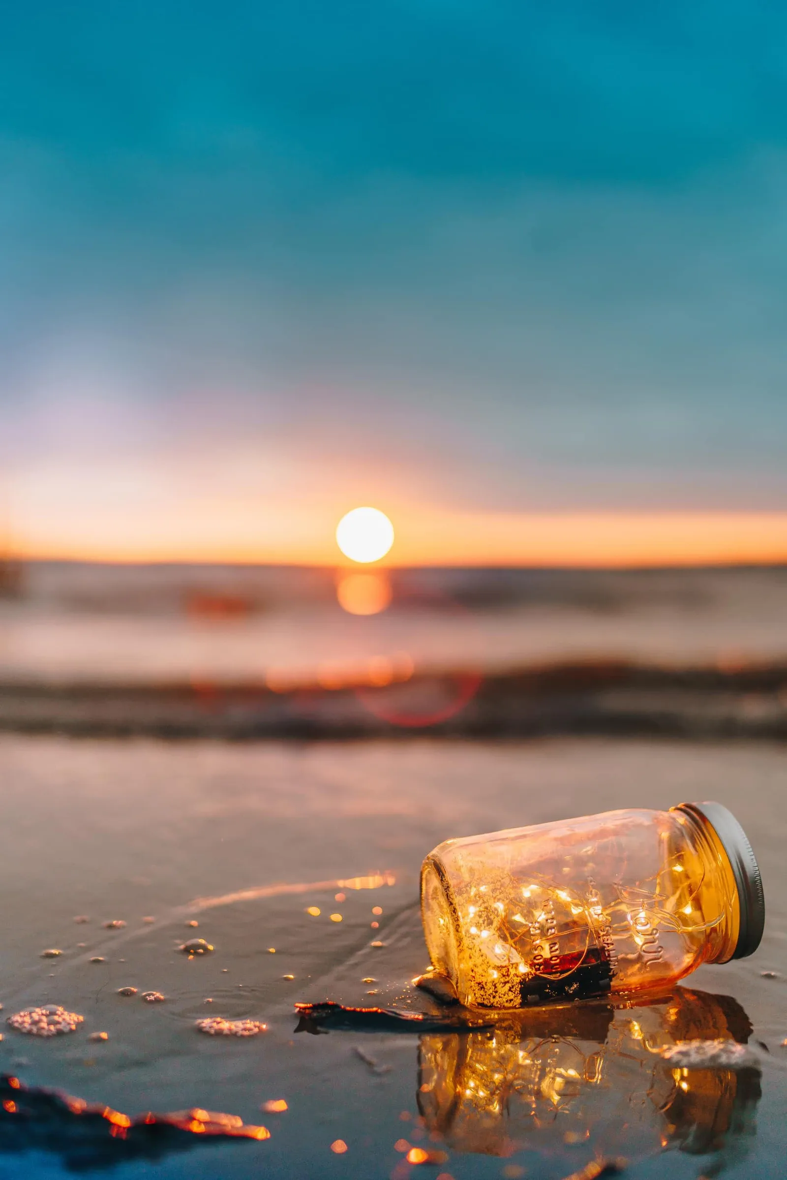 Sonnenuntergang über Meer mit Glaskrug gefüllt mit leuchtenden Partikeln am Strand