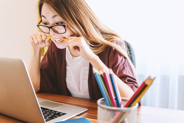 Frau mit Brille sitzt am Laptop und hält Bleistift im Mund, fröhlich