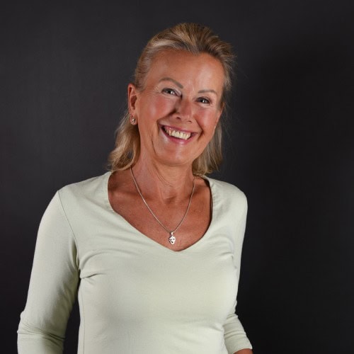 Marina Tscheschel, blonde Frau mit freundlichem Lächeln in cremefarbenem Oberteil vor dunklem Hintergrund
