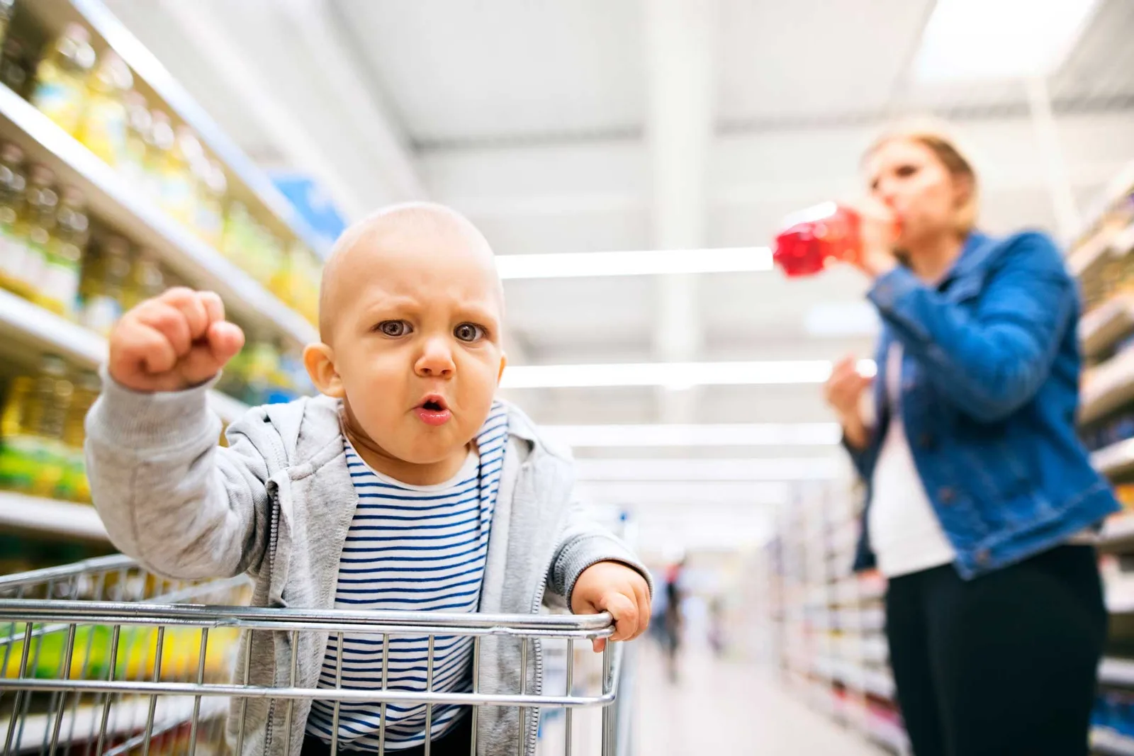 Kleinkind im Supermarkt-Einkaufswagen bei Trotzanfall mit Mutter im Hintergrund