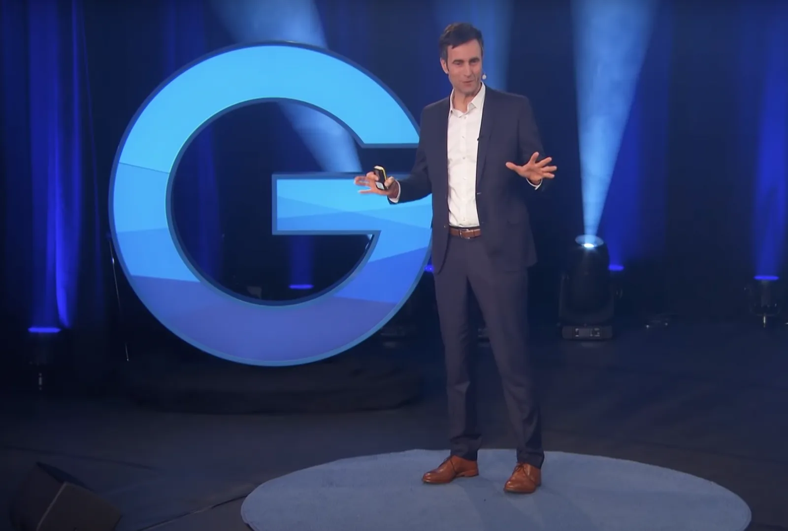 Gesprächsführung – Videokurs für Führungskräfte von Markus Jotzo 2 Markus Jotzo präsentiert auf einer Bühne vor großem blauen G-Logo