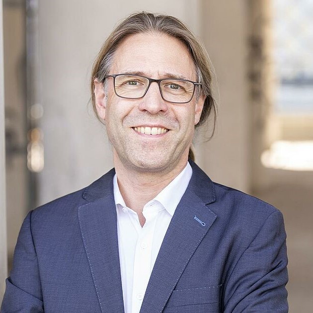 Porträt Franz Sperlich, Mann mit Brille im blauen Blazer vor hellem Hintergrund