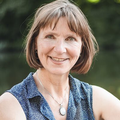 Porträtfoto von Anita Maas, Frau mit braunen Haaren und blauem Oberteil vor grünem Hintergrund