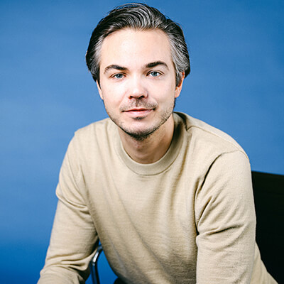 Porträtfoto von Timo, junger Mann mit dunklen Haaren und Beige-Shirt vor blauem Hintergrund