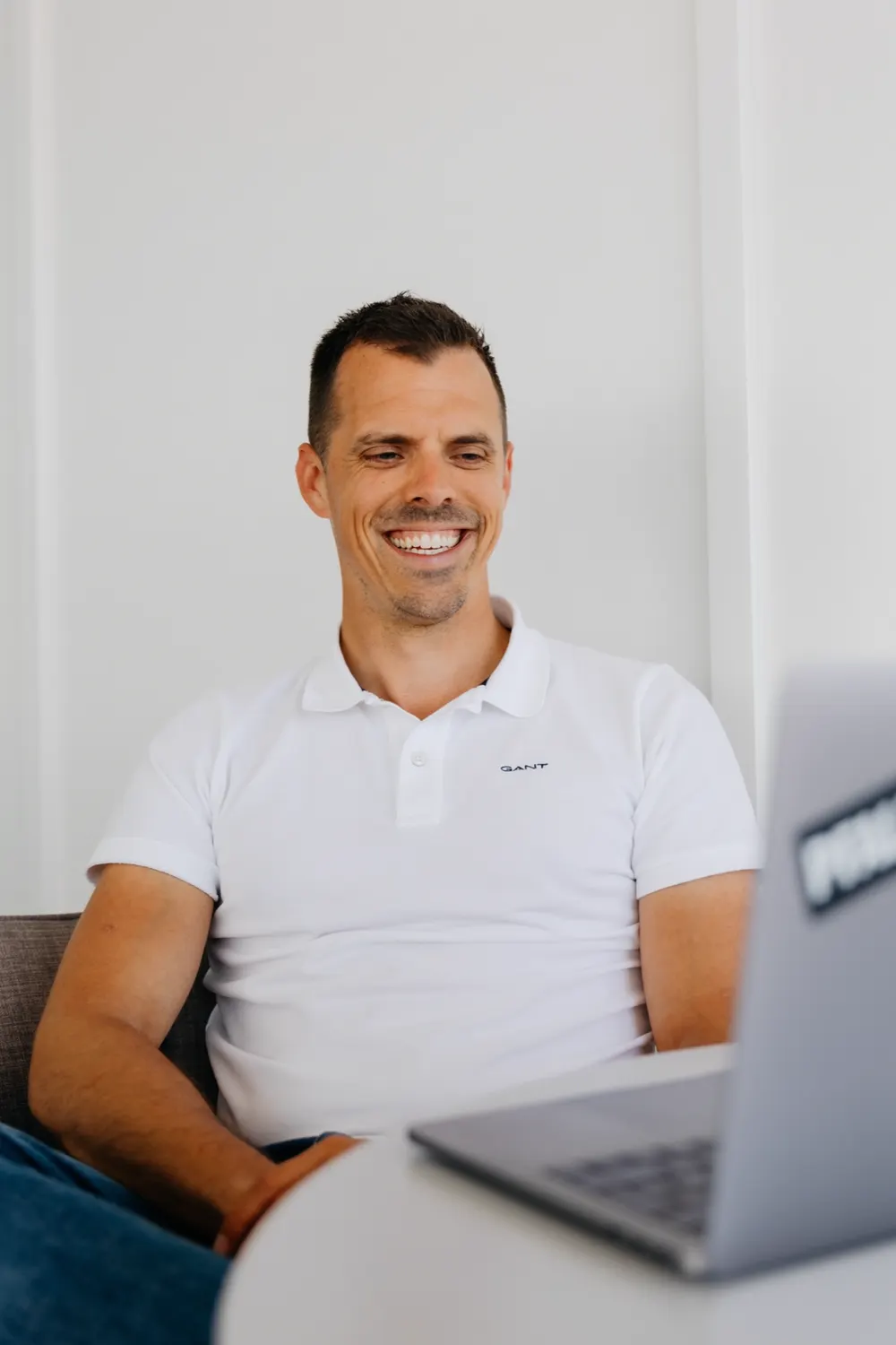 Mann in weißem Poloshirt lacht vor Laptop mit unscharfem Hintergrund