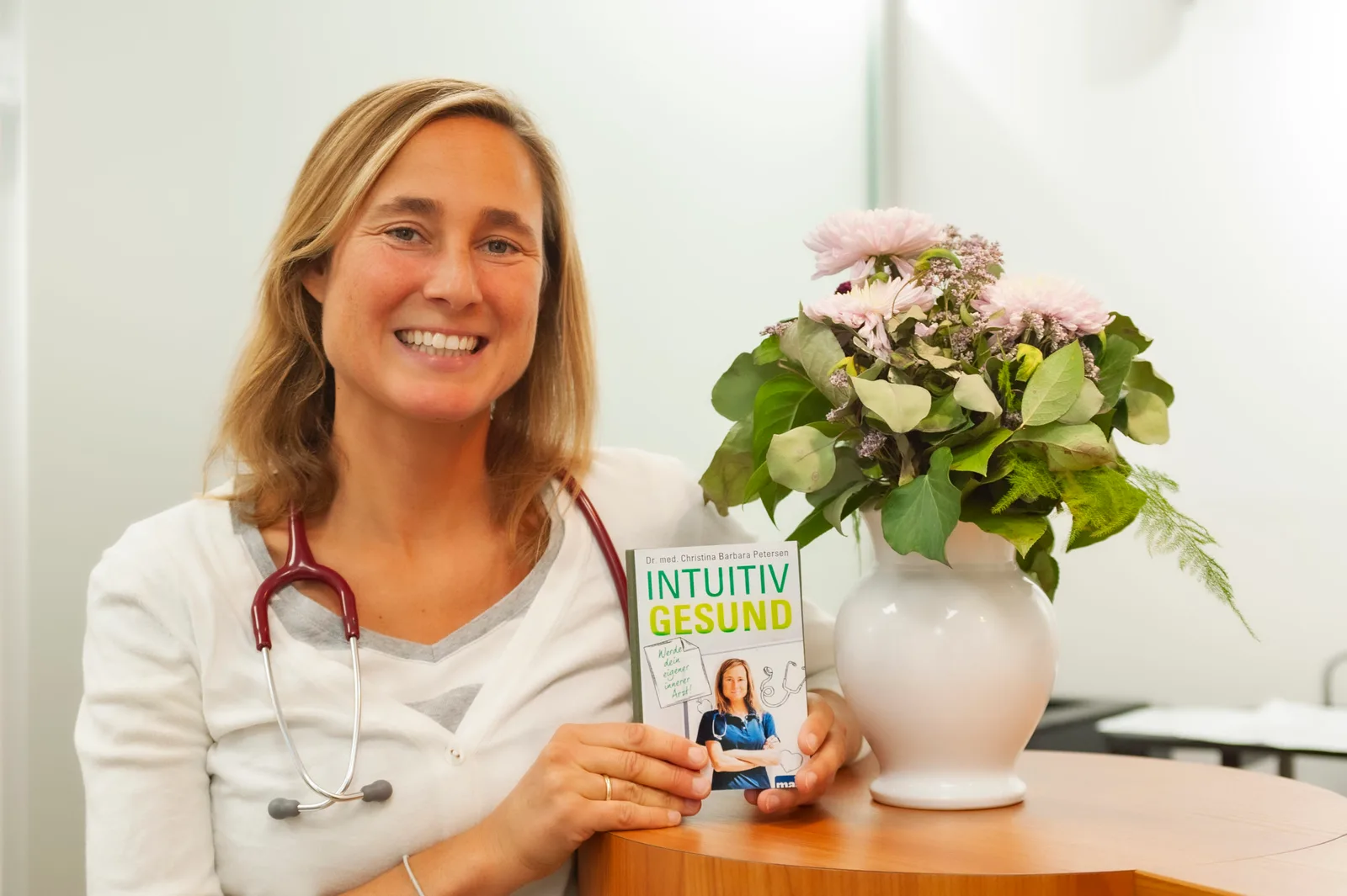 Dr. med. Christina Petersen mit Stethoskop hält Buch Intuitiv Gesund in hellem Büro mit Blumengesteck