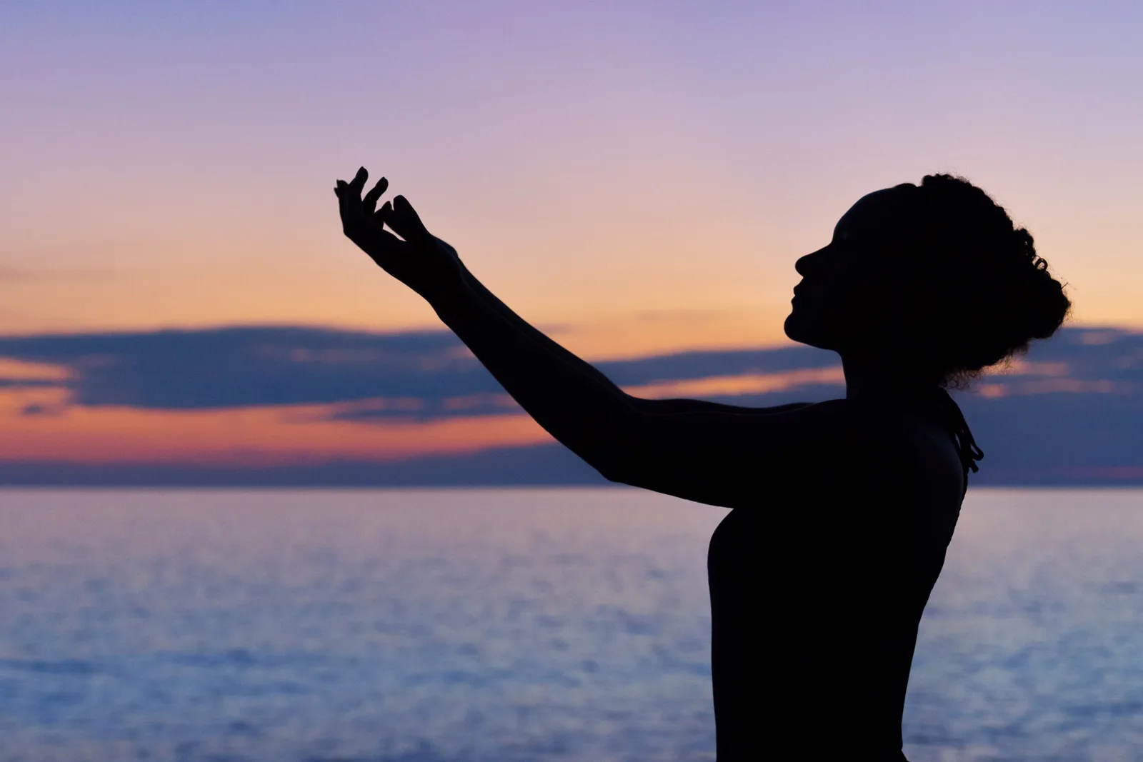 Silhouette einer Frau bei Sonnenuntergang am Meer, Arm ausgestreckt
