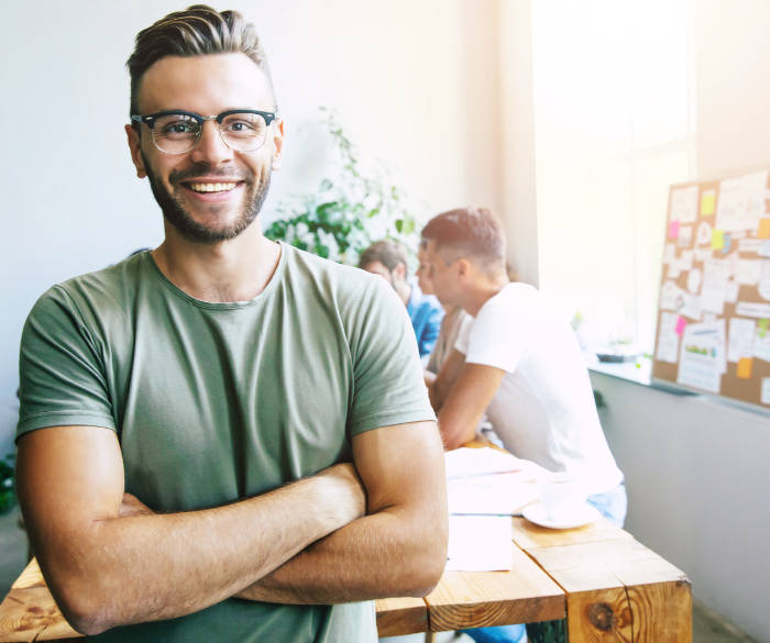Junger Mann in grünem Shirt lächelt, im Hintergrund Workspace mit Kollegen und Pinnwand