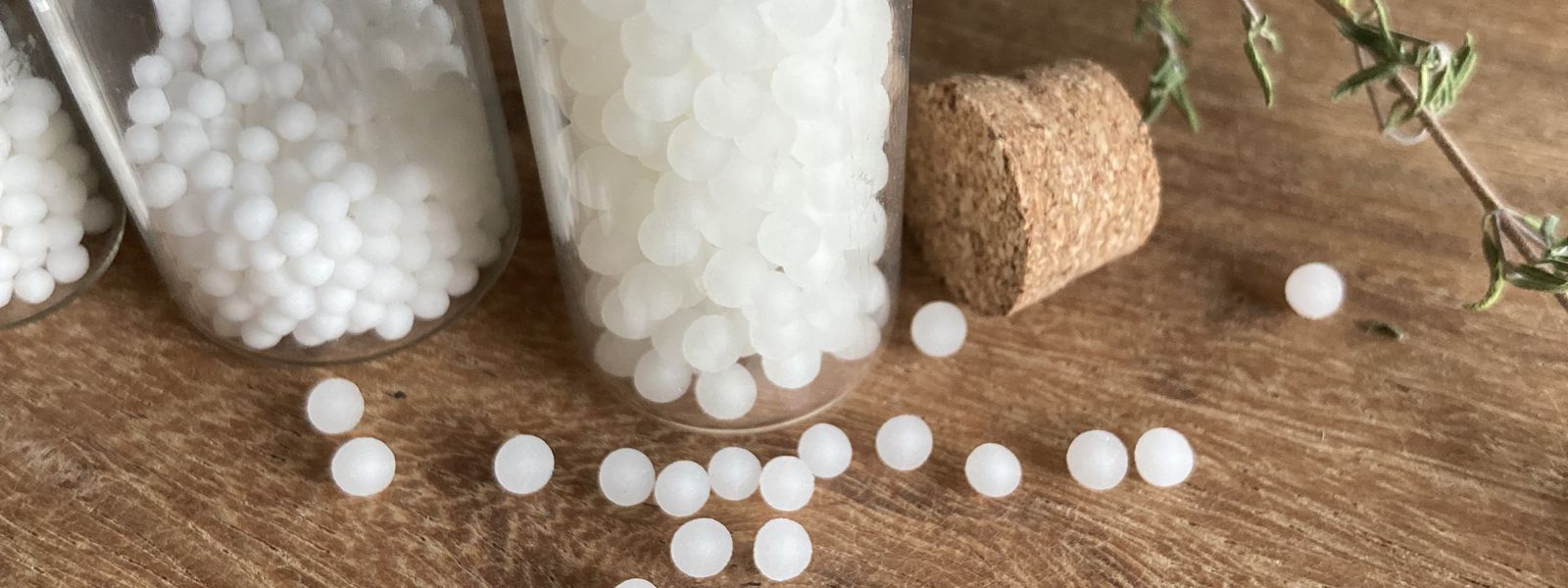 Homöopathische Globuli in Gläsern und verstreut auf Holztisch mit Pflanzenmaterial