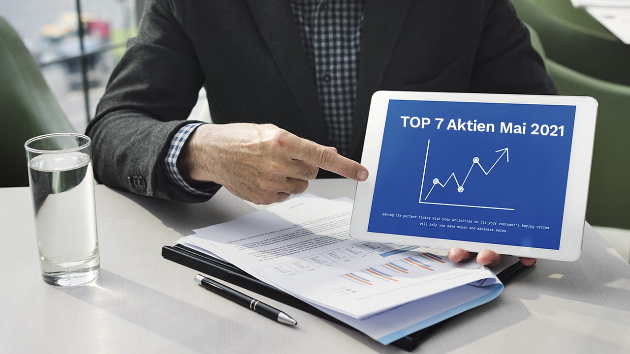 Mann zeigt auf Tablet mit 'TOP 7 Aktien Mai 2021' Grafik, Wasserglas auf Tisch