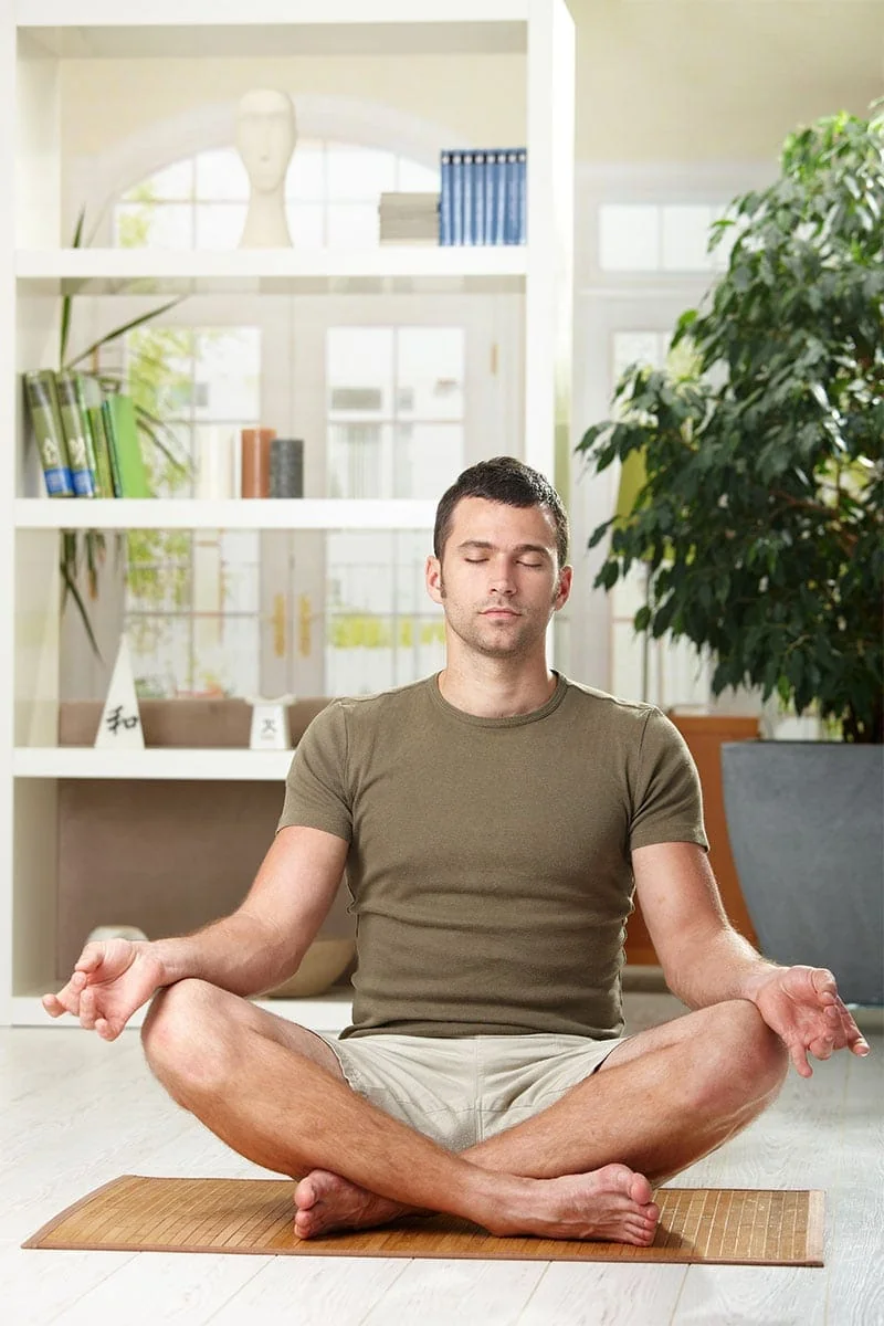 Mann sitzt meditierend auf Yogamatte im hellen Wohnzimmer