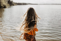 Frau von hinten am See stehend, braune Bohemian-Kleidung, Wald im Hintergrund