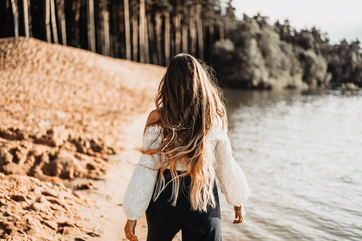 Sarah Jane Rogalski am Seeufer, von hinten, lange Haare, weiße Strickjacke über schwarzem Oberteil