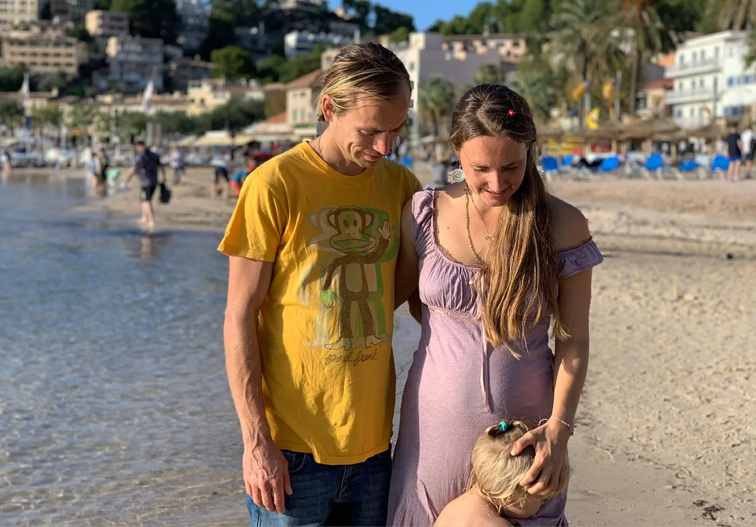 Junges Paar am Strand spazierend, Mann in gelbem Shirt, Frau in lilafarbenem Kleid mit kleinem Hund