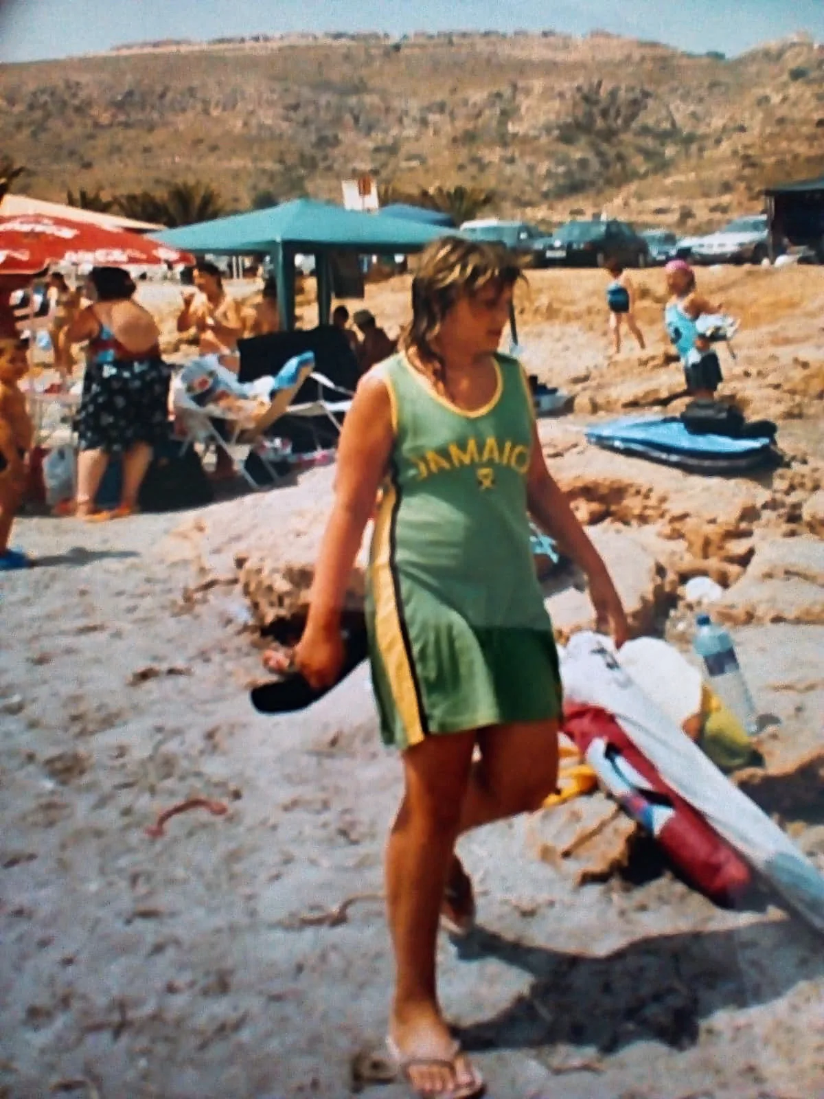 Junge in grünem Badeanzug am Strand mit Surfboard im Hintergrund