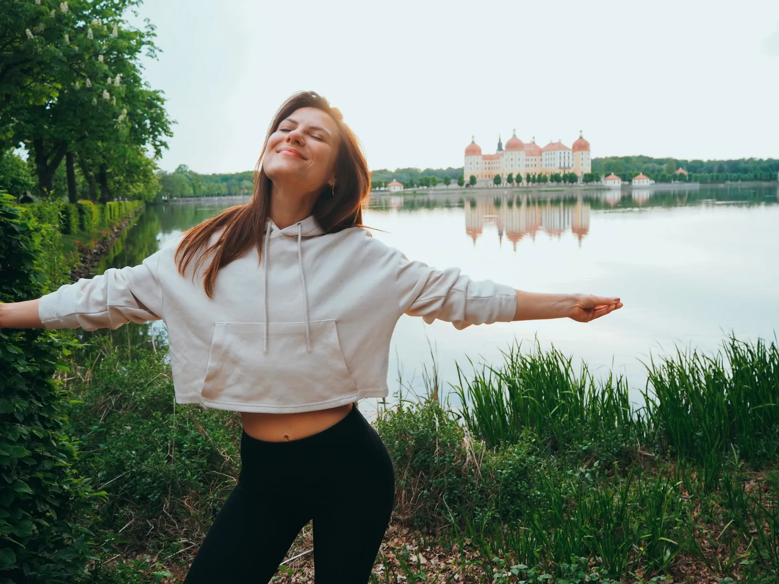 Frau in weißer Sportjacke mit offenen Armen am See vor Schloss
