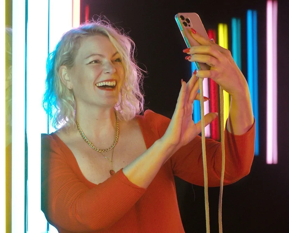 JyotiMa Flak hält lachend Smartphone mit bunten Neon-Stäben im Hintergrund