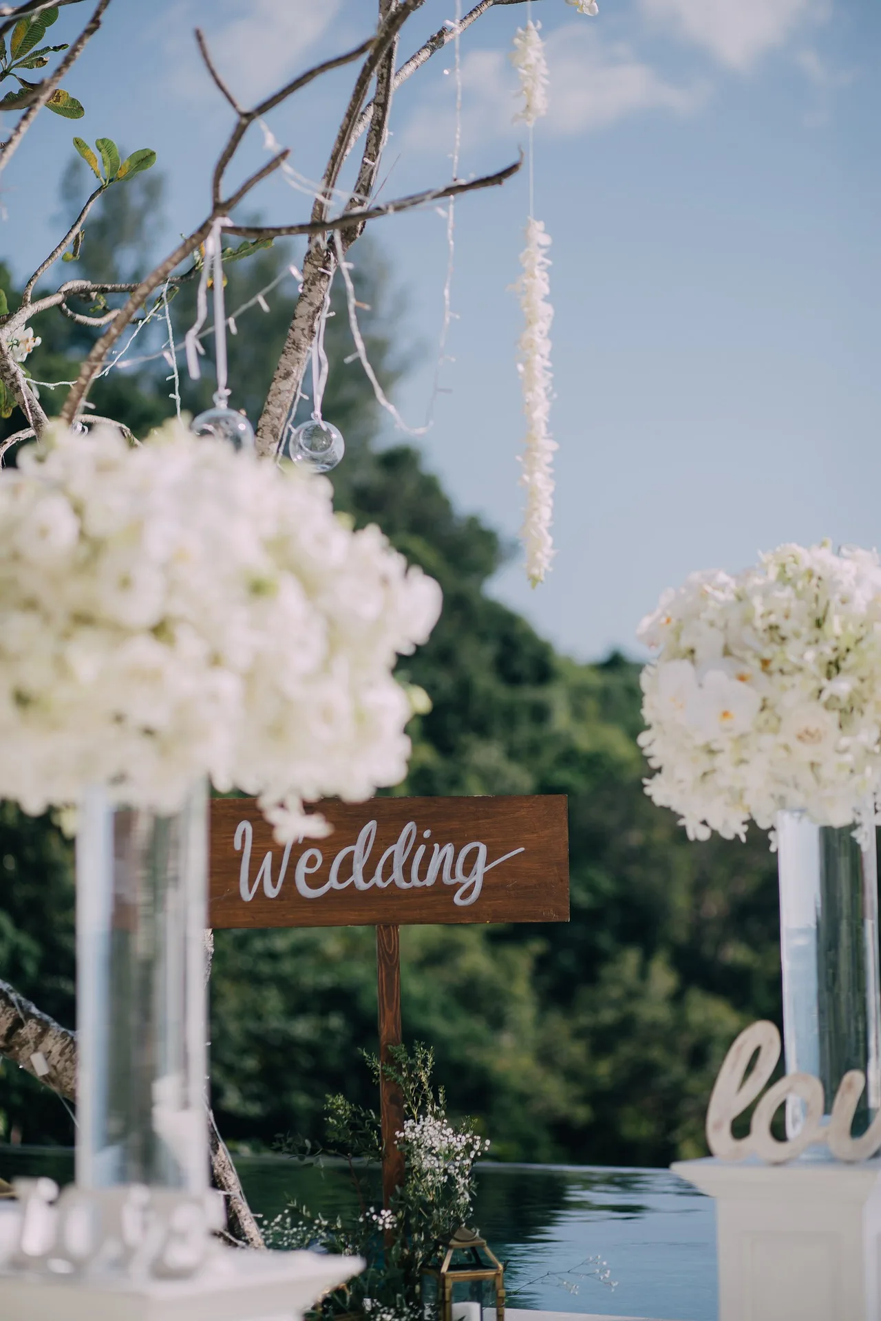 Hochzeitsdekoration im Freien mit weißen Hortensien an Ästen, hängenden Glaskugeln und Holzschild mit Aufschrift Wedding