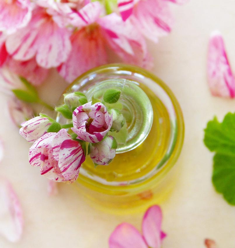 Ölglas mit rosafarbener Blüte und grünem Öl vor unscharfem Hintergrund