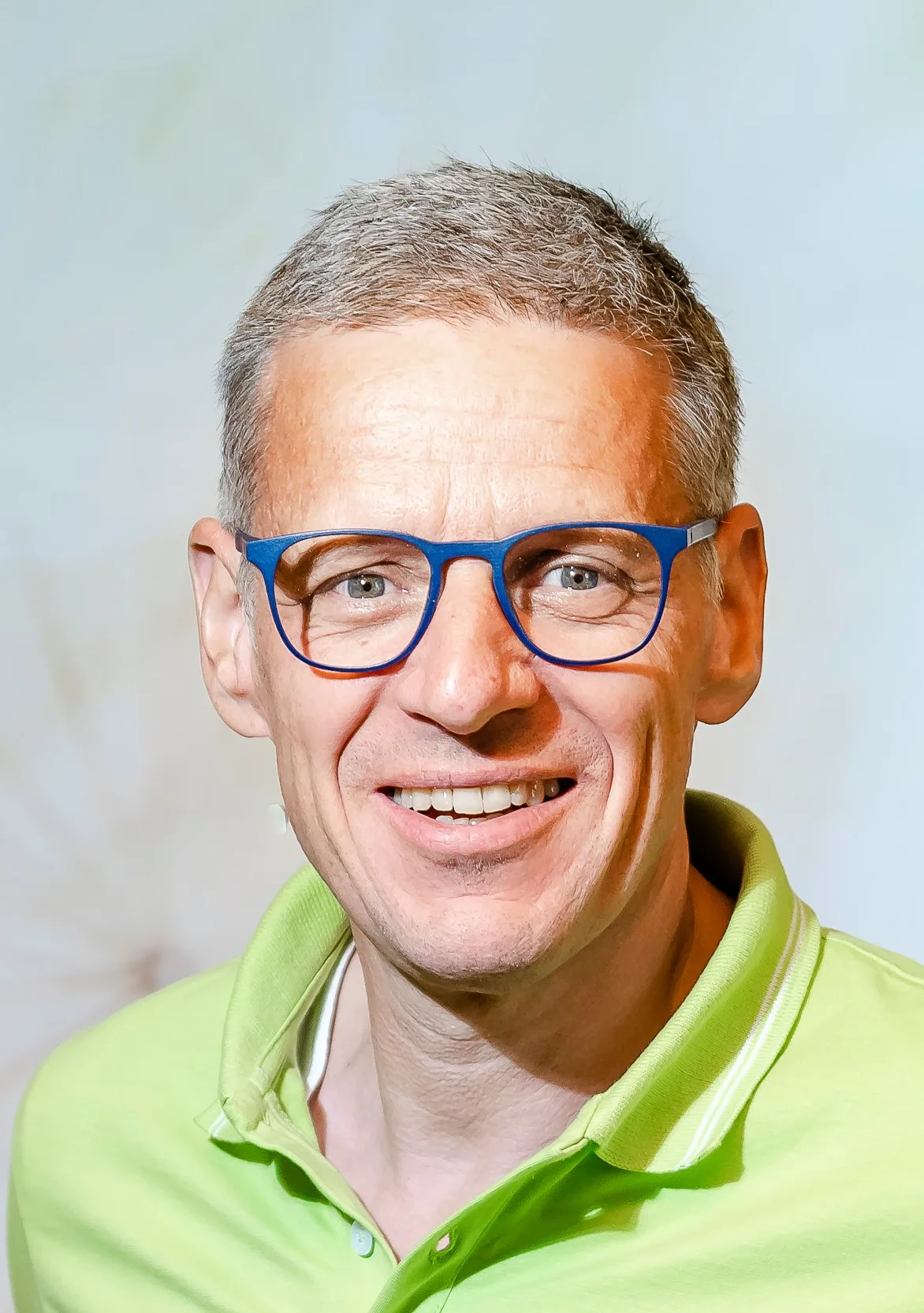 Mann mit kurzen graubraun Haaren, blauer Brille mit oranger Einfassung, grünem Poloshirt, lächelt in Kamera