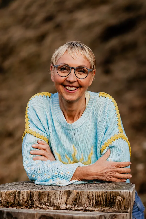 Frau mit blondem Kurzschnitt, Brille, hellblauem Strickpulli mit gelben Details, lehnt an Holzklotz vor Naturhintergrund