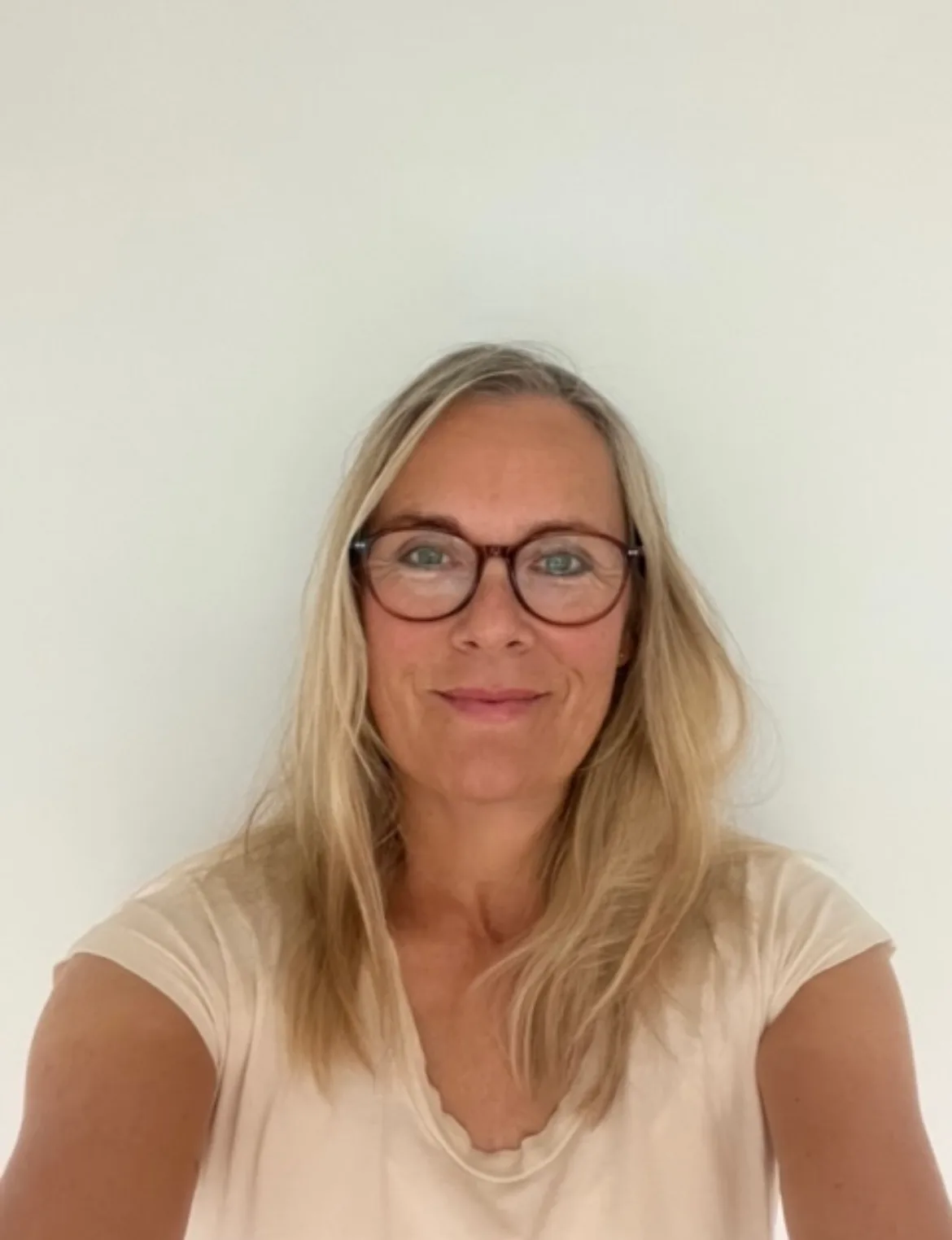 Frau mit langem blondem Haar, Brille mit braunem Rahmen, helles T-Shirt, neutral lächelnd vor weißem Hintergrund