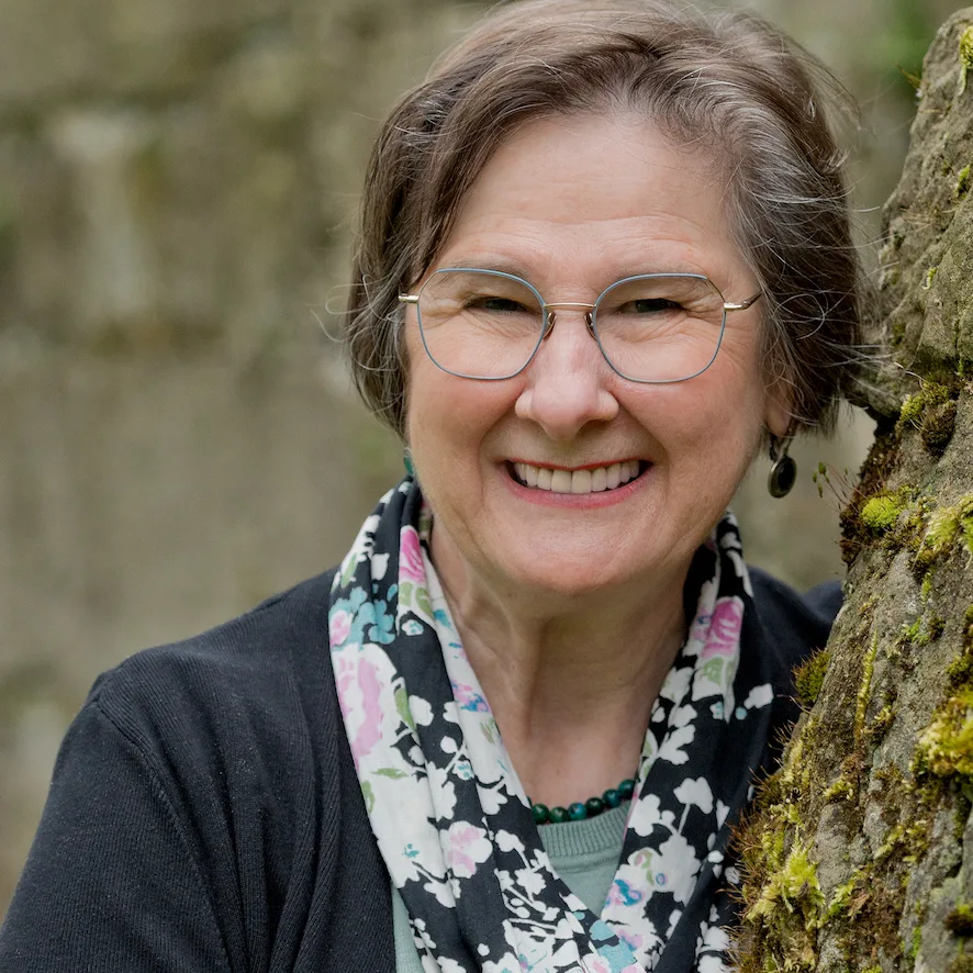 Frau mittleren Alters mit braunen Haaren, grauer Brille, buntem Blumen-Schal, schwarzer Jacke, lächelt neben moosigem Baumsta