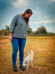 Frau in grauem Shirt und blauer Hose steht mit weißem kleinem Hund auf freiem Feld