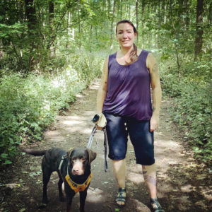 Frau mit schwarzem Hund in gelber Leine auf Waldweg spazierend