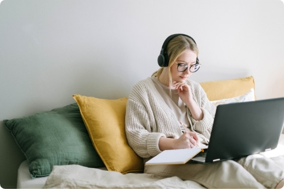 Junge Frau mit Kopfhörern und Brille sitzt auf Couch mit Laptop