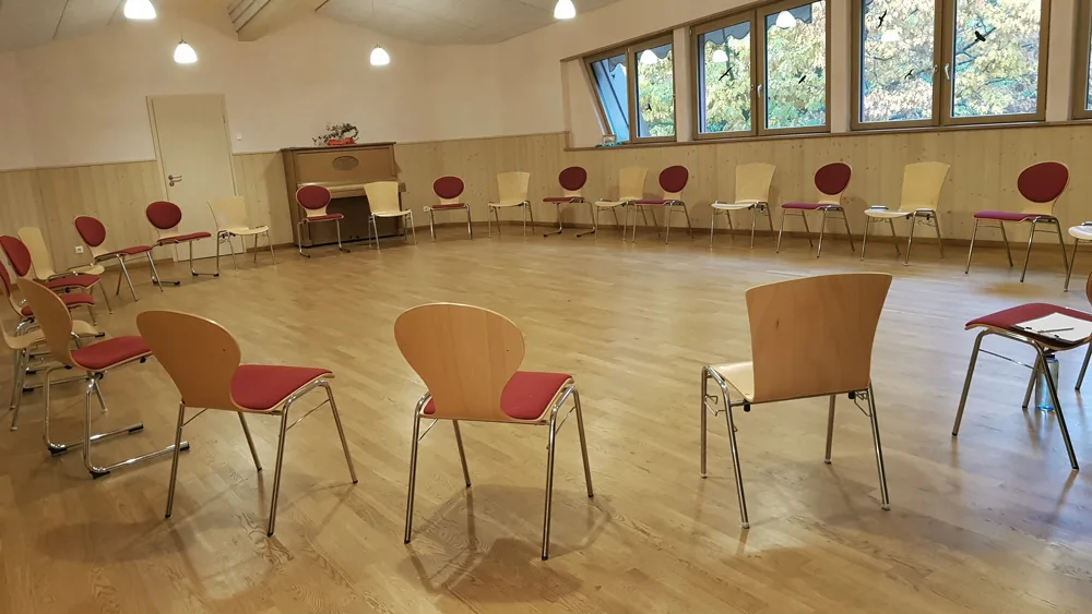 Leerer Aufstellungsraum mit Holzboden und Stühlen mit roten Kissen in Halbkreis-Anordnung, helle Fenster