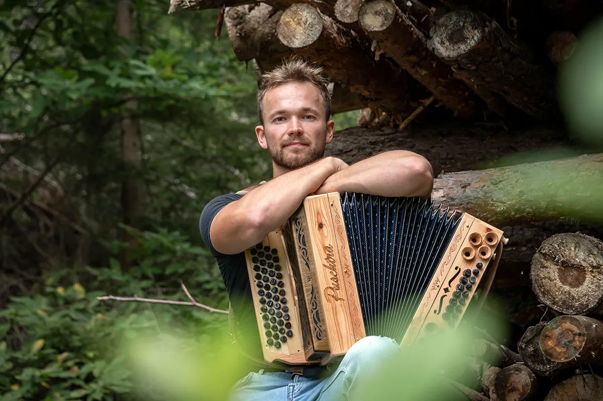 Harmonikalehrer Manuel Merler mit Harmonika im Freien vor Holzstapel
