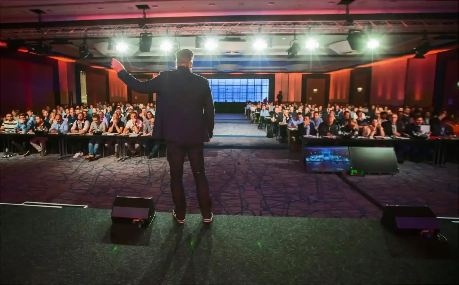 Mann auf Bühne vor großem Publikum in beleuchteter Konferenzhalle, spricht zu Zuhörern