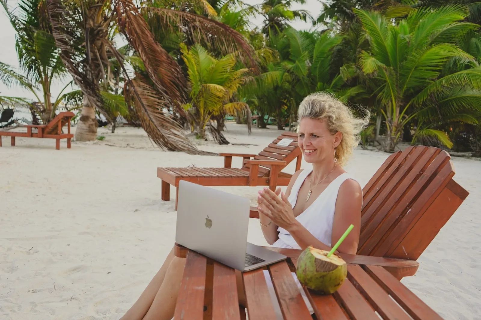 Blonde Frau mit rosa Muschel-Accessoire am Laptop im Strandstühle mit Kokosgetränk