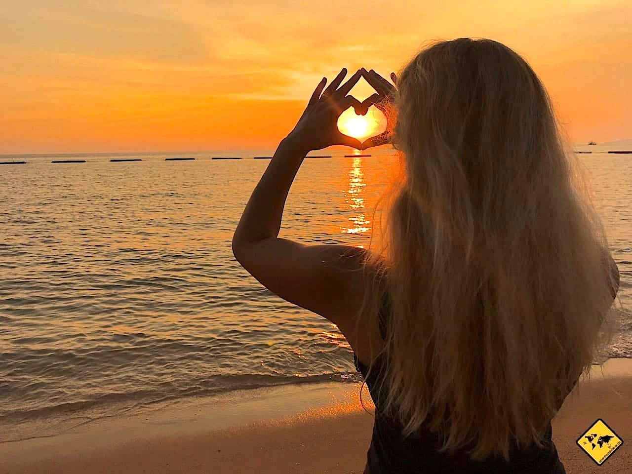 Frau mit langen Haaren macht Herzzeichen mit Händen vor Sonnenuntergang über Meer